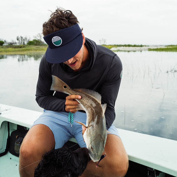 Man holding a fish on a boat with a visor and jacket, surrounded by water and greenery.