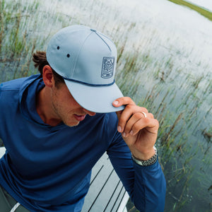 Man wearing a light blue cap with a logo, standing by a body of water.