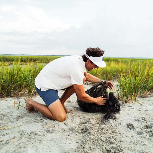Person kneeling on a sandy beach with a dog, surrounded by grass and water.