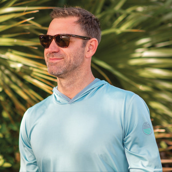 Man wearing sunglasses and a light blue shirt in front of green foliage